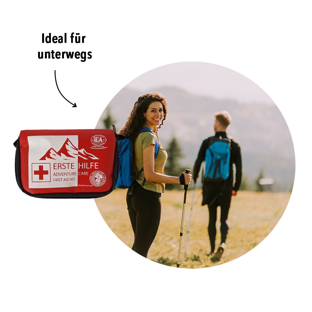 Personen wandern mit Rucksäcken. Im Vordergrund eine rote Erste-Hilfe-Tasche.