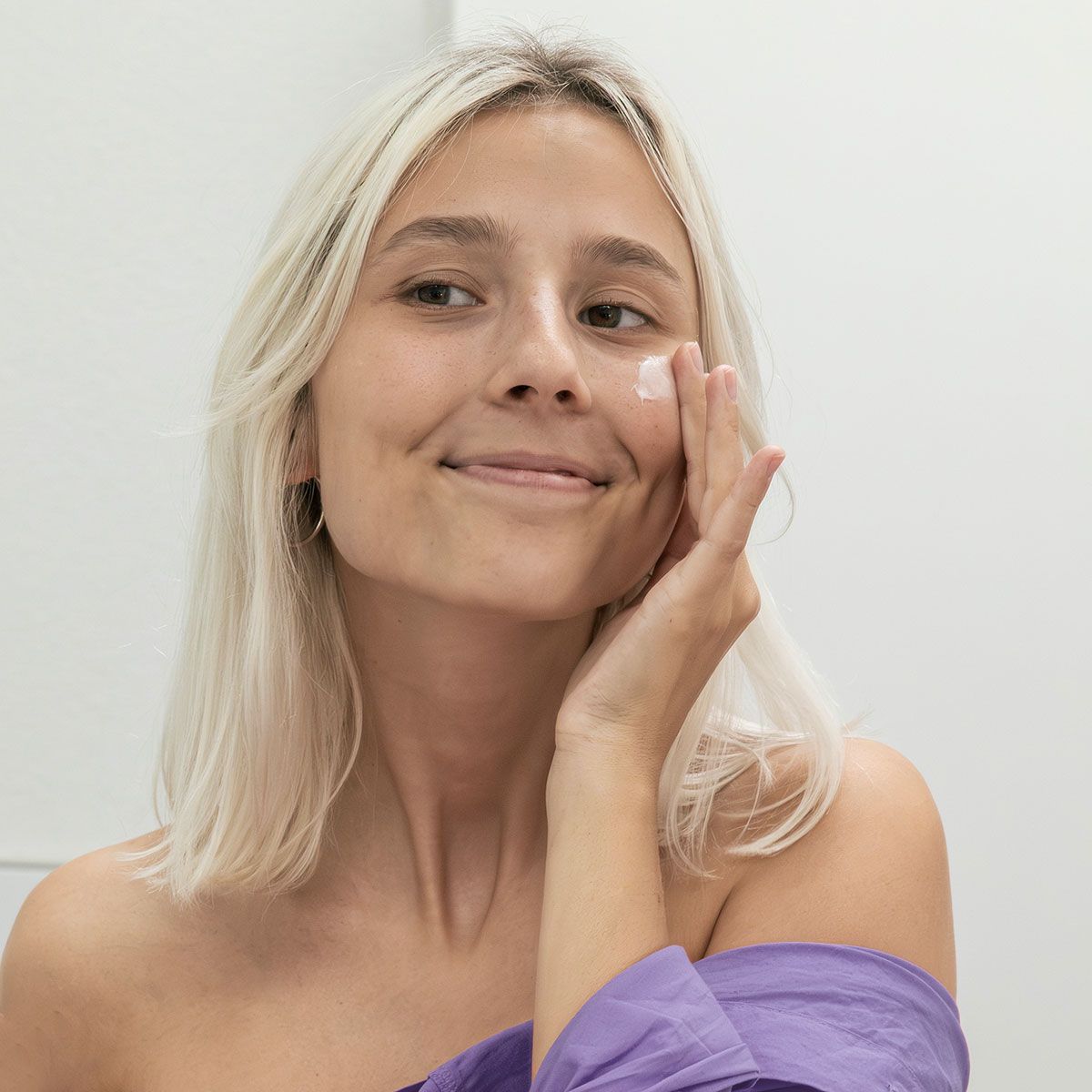 Frau trägt Creme auf ihr Gesicht auf. Sie lächelt. Helle Haare, lila Oberteil.