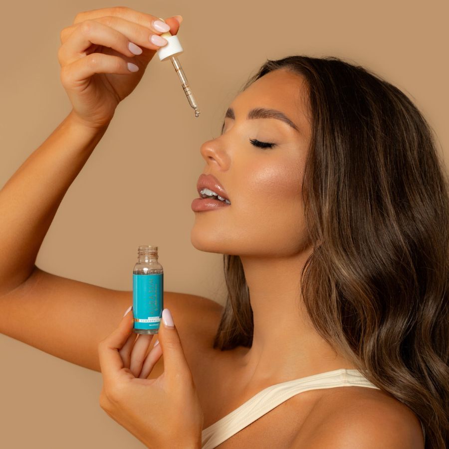 Eine Frau hält eine kleine Glasflasche mit blauem Etikett und einen Tropfer mit Serum über ihrem Gesicht.