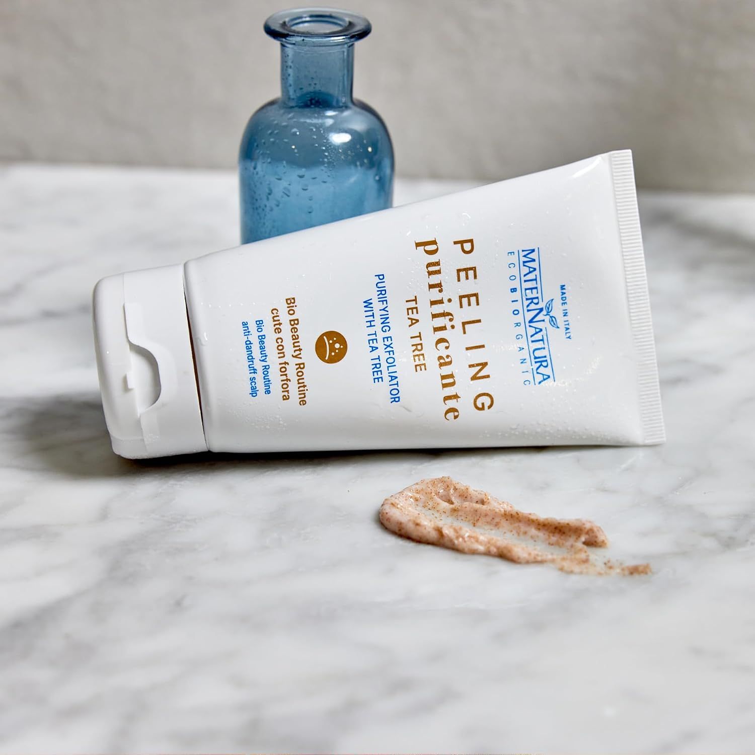 Weißer Spender mit Peeling und Teebaumöl. Ein kleiner blauer Flakon steht dahinter.
