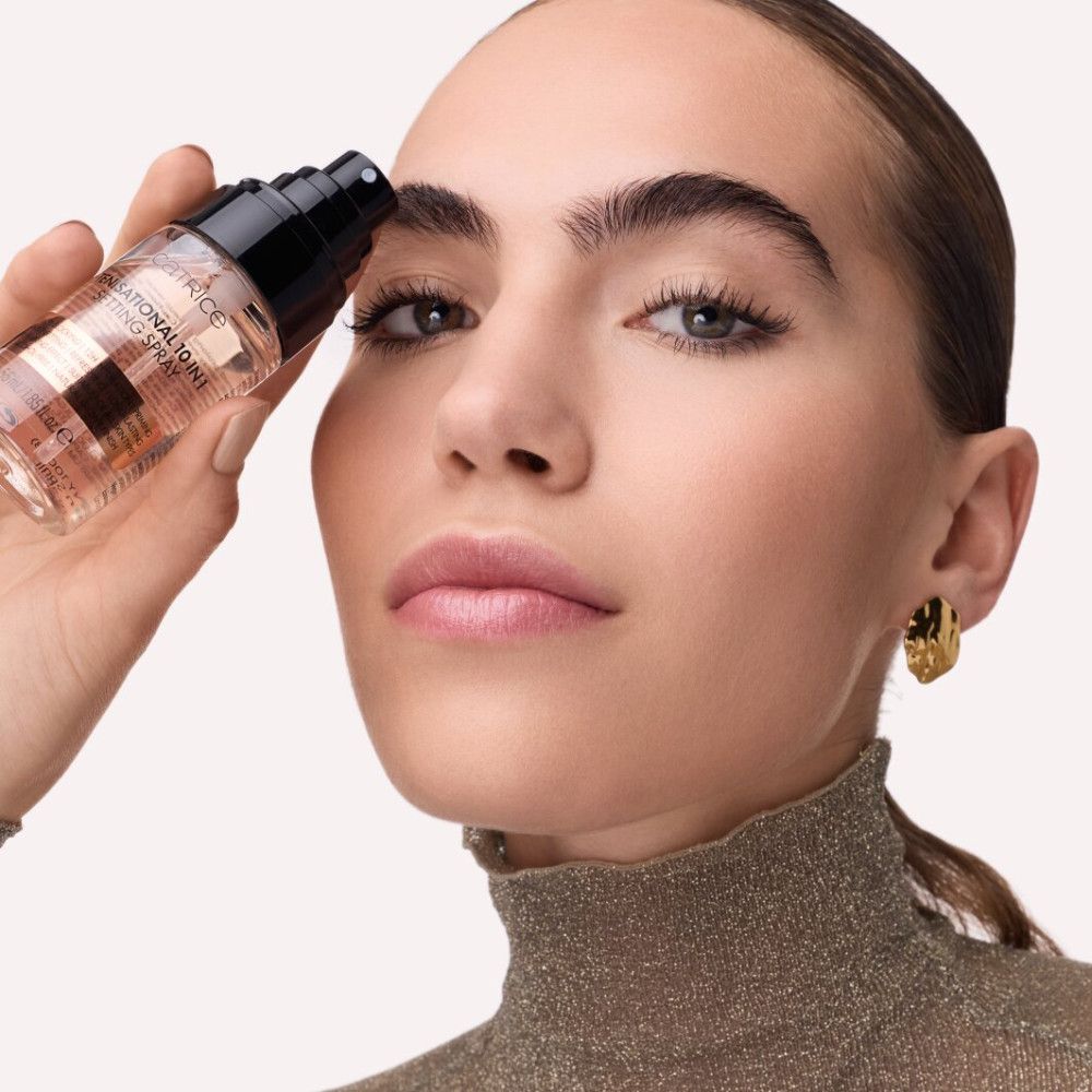 Frau hält Setting Spray vor Gesicht. Produktname und Eigenschaften sichtbar. Goldfarbener Kragen und Ohrringe.