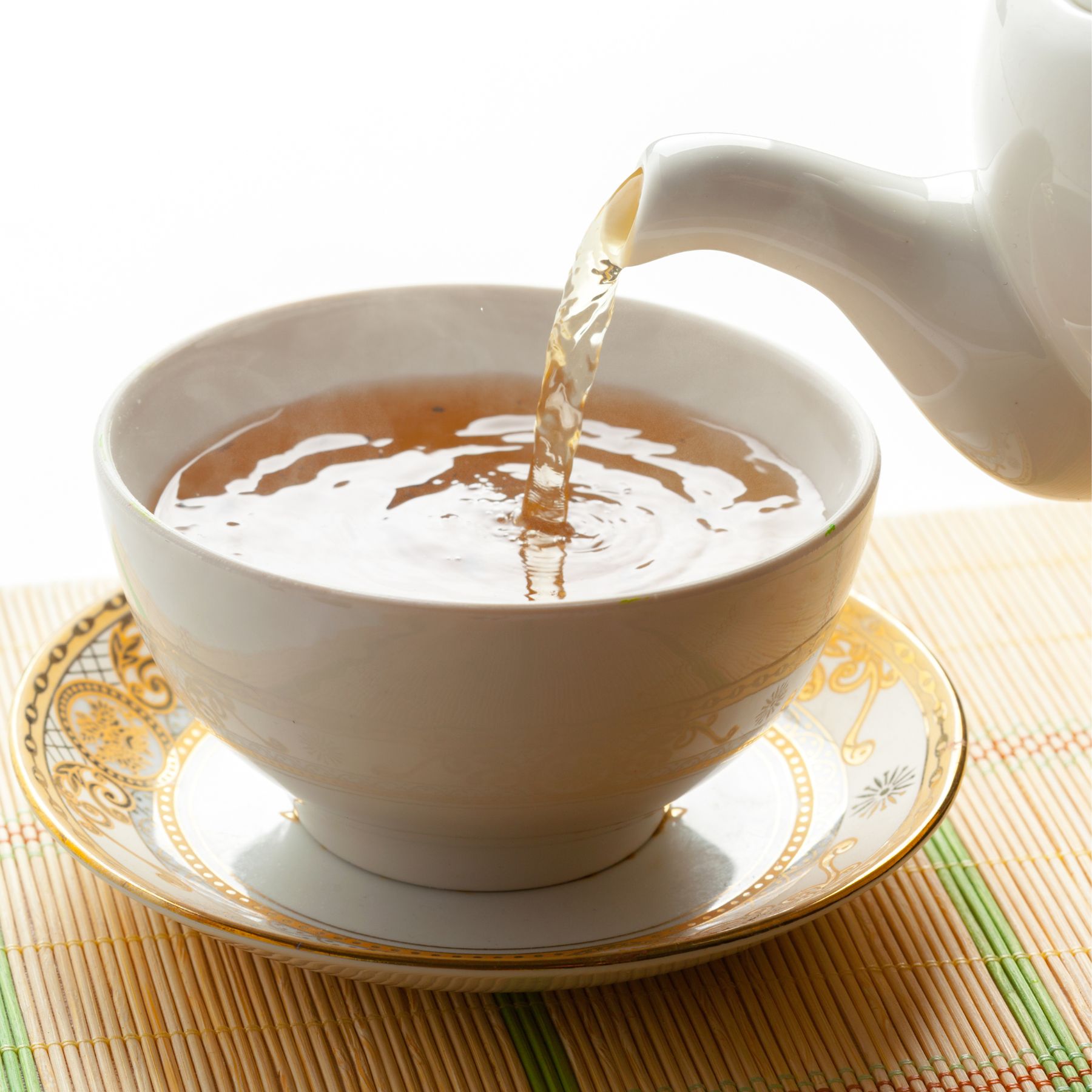 Weißer Tee wird aus einer weißen Kanne in eine weiße Tasse gegossen. Untertasse mit goldenem Muster. Auf Bambusmatte.