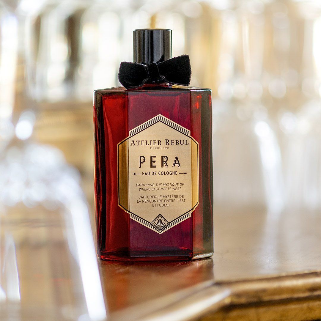 Rote Glasflasche mit schwarzem Verschluss und Schleife. Aufschrift: Atelier Rebul Pera Eau de Cologne. Auf einem Tisch platziert.