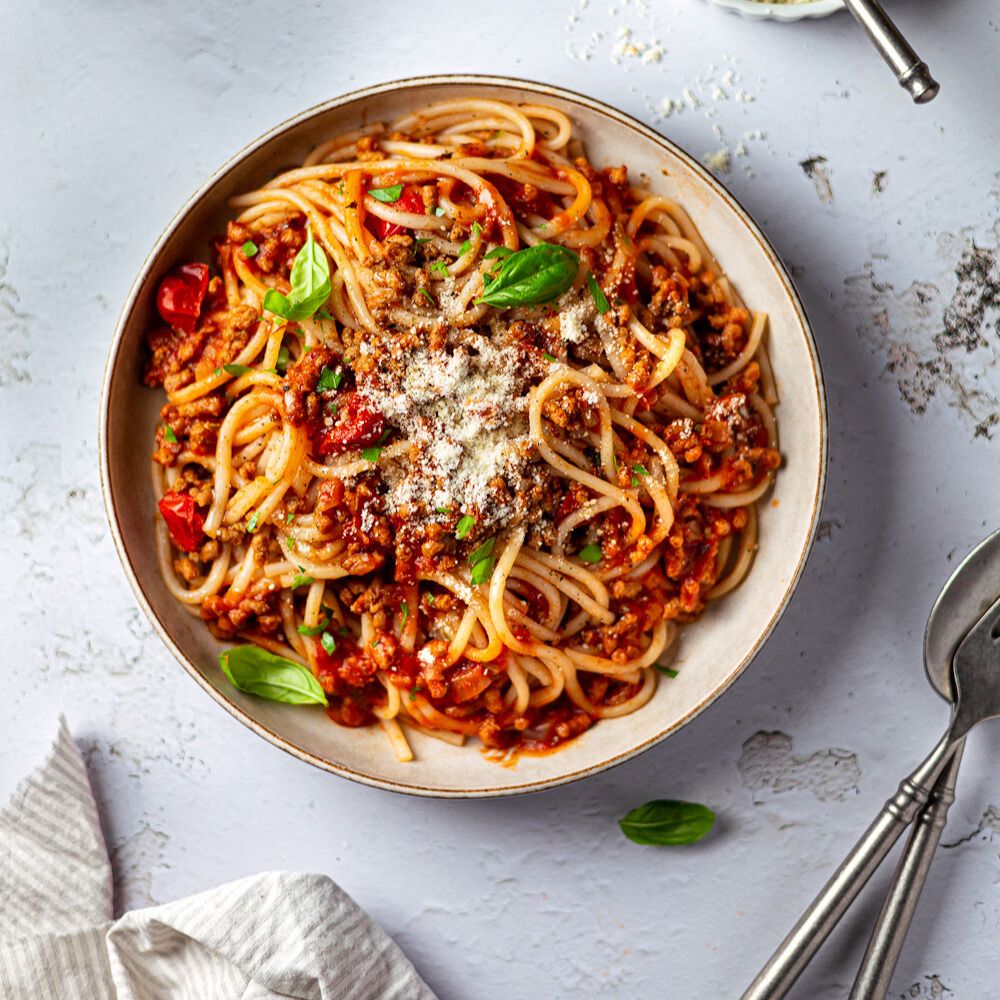 Teller mit Spaghetti mit Tomaten, Fleisch, Käse und Basilikum. Gabel und Löffel daneben. Auf dem Tisch: Serviette.
