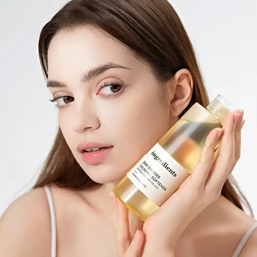 Frau hält eine Flasche ONGREDIENTS Skin Barrier Calming Softener. Produktetikett deutlich sichtbar. Frau blickt in die Kamera.