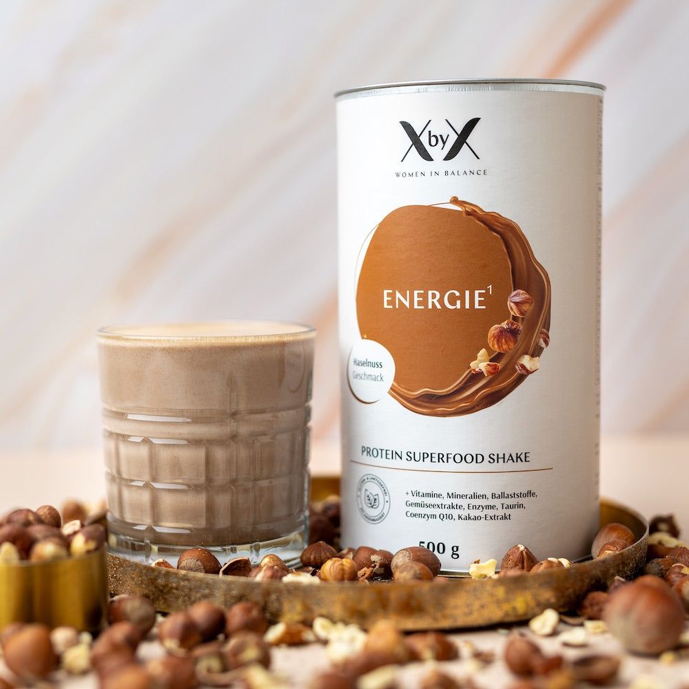 Dose und Glas mit braunem Getränk. Dose mit Aufschrift 'Energie', 'Protein Superfood Shake'. Haselnüsse umgeben das Glas.