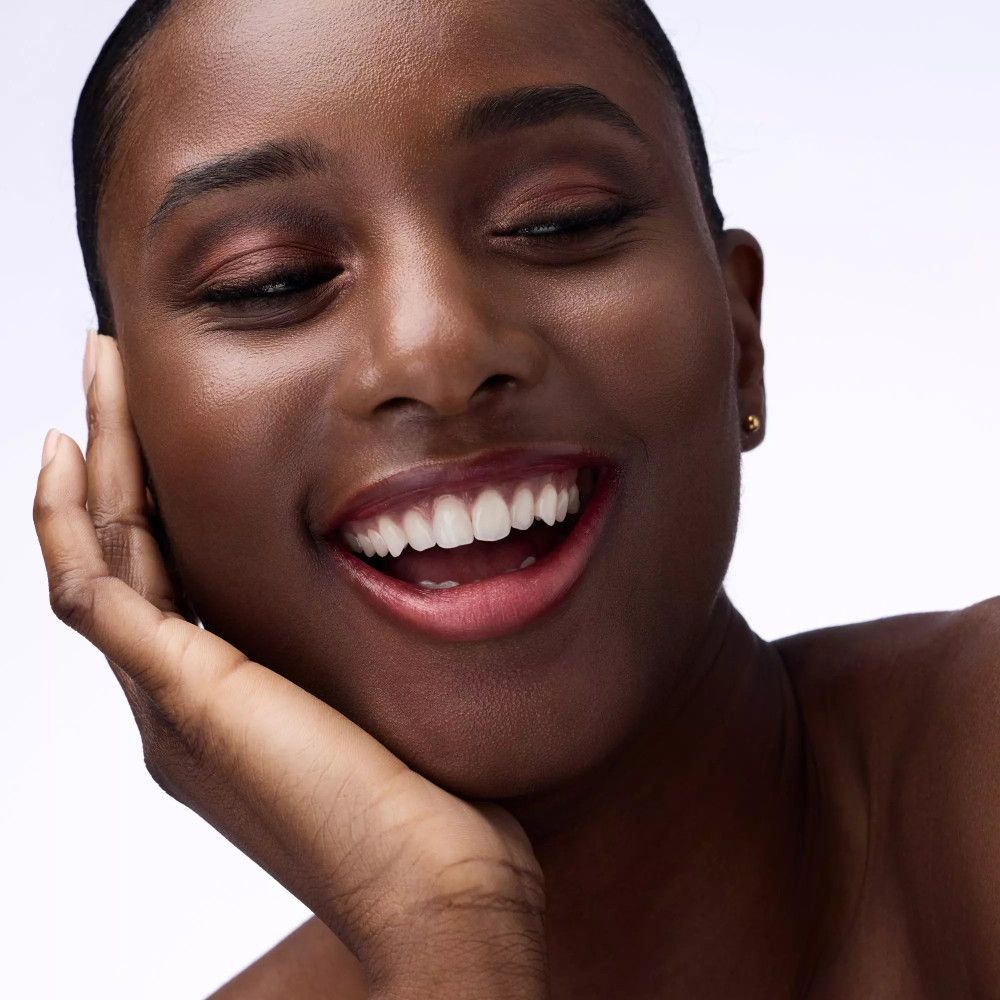 Porträt einer lächelnden Frau mit Lidschatten. Farben: Orange und Braun. Hautton: dunkel. Sie berührt ihr Kinn.