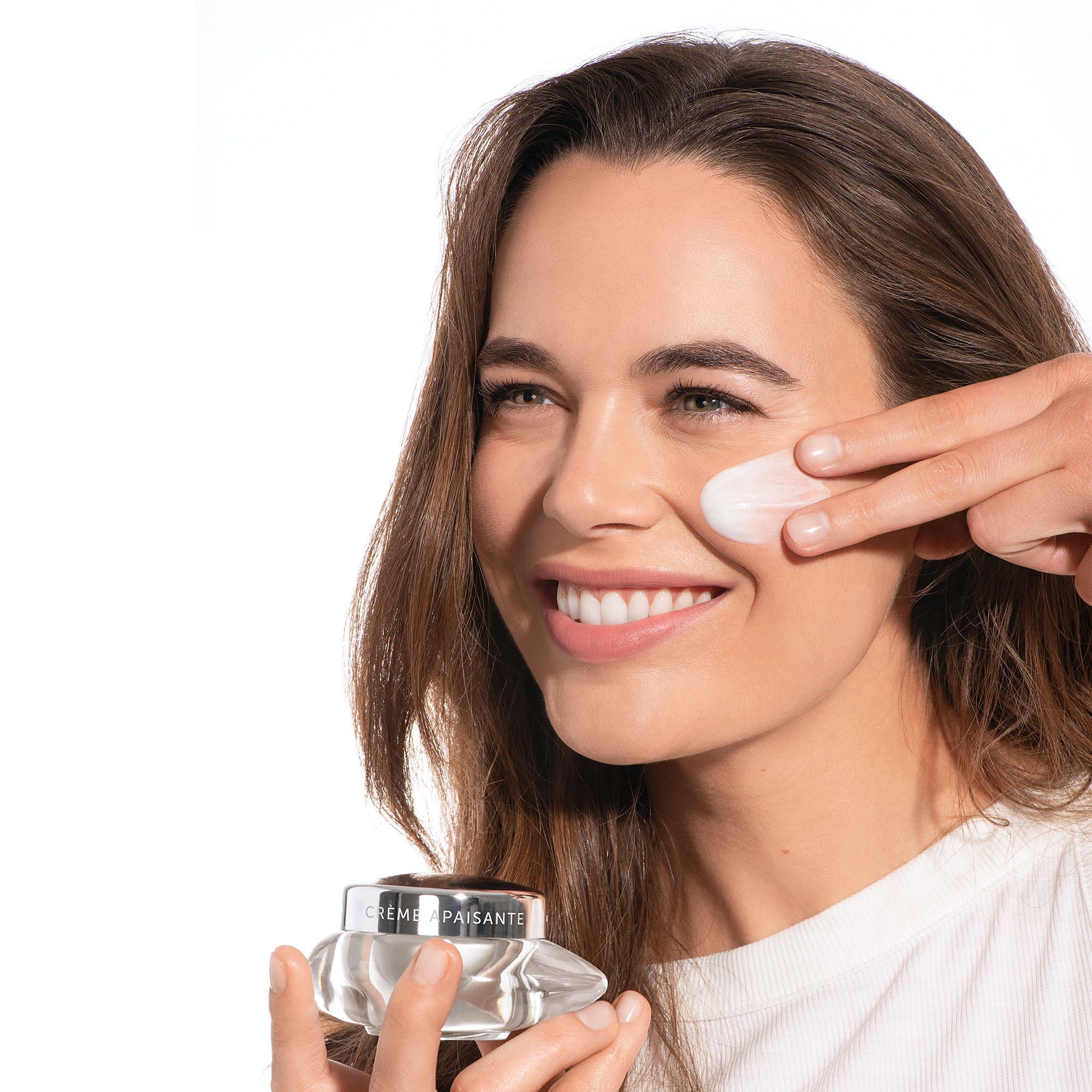 Frau trägt Creme auf. Glasbehälter mit silbernem Deckel. Aufschrift: CRÈME APAISANTE.