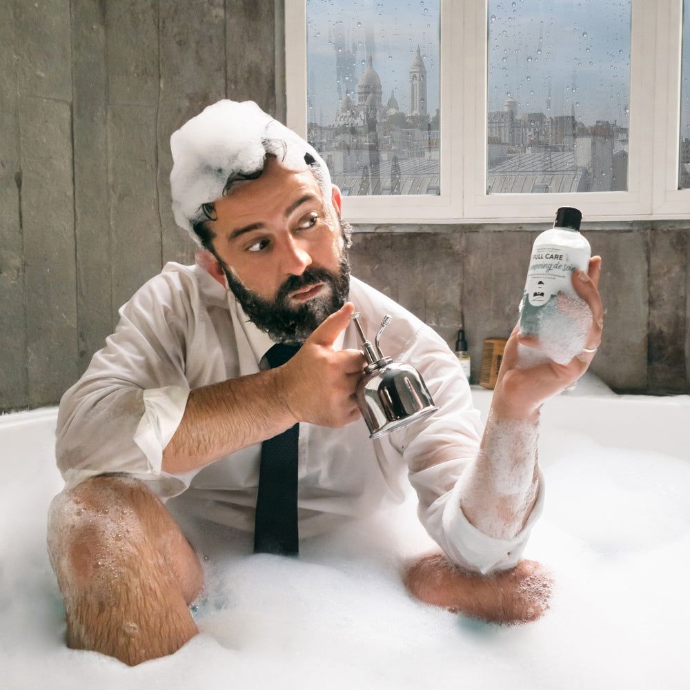 Mann im Anzug in Badewanne mit Schaum. Hält eine Flasche Monsieur BARBIER 2-in-1 Bart- und Haarshampoo.