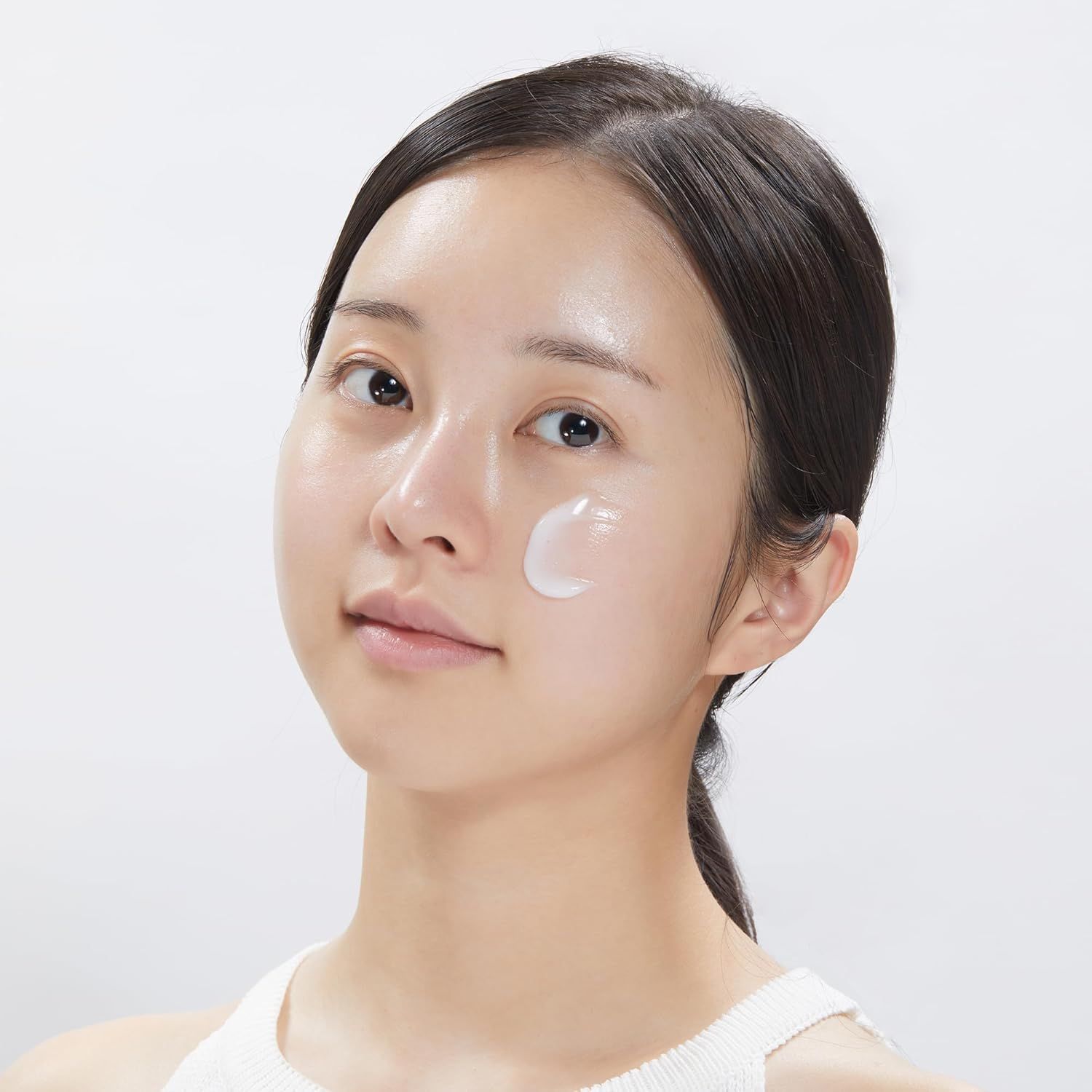 Nahaufnahme einer Frau, die Creme auf ihr Gesicht aufträgt. Helle Haut, dunkle Haare. Weißes Tanktop.