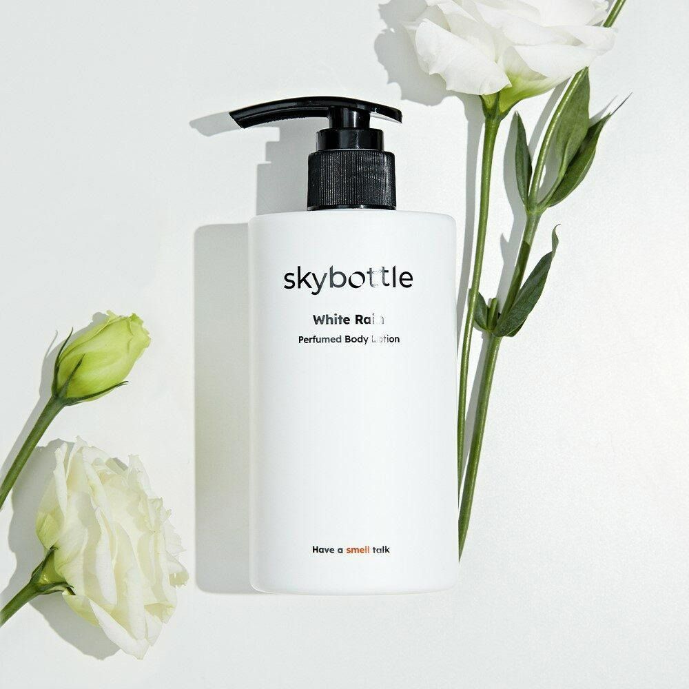 Weiße SKYBOTTLE-Körperlotion mit schwarzem Pumpkopf, umgeben von weißen Blüten und grünen Blättern.