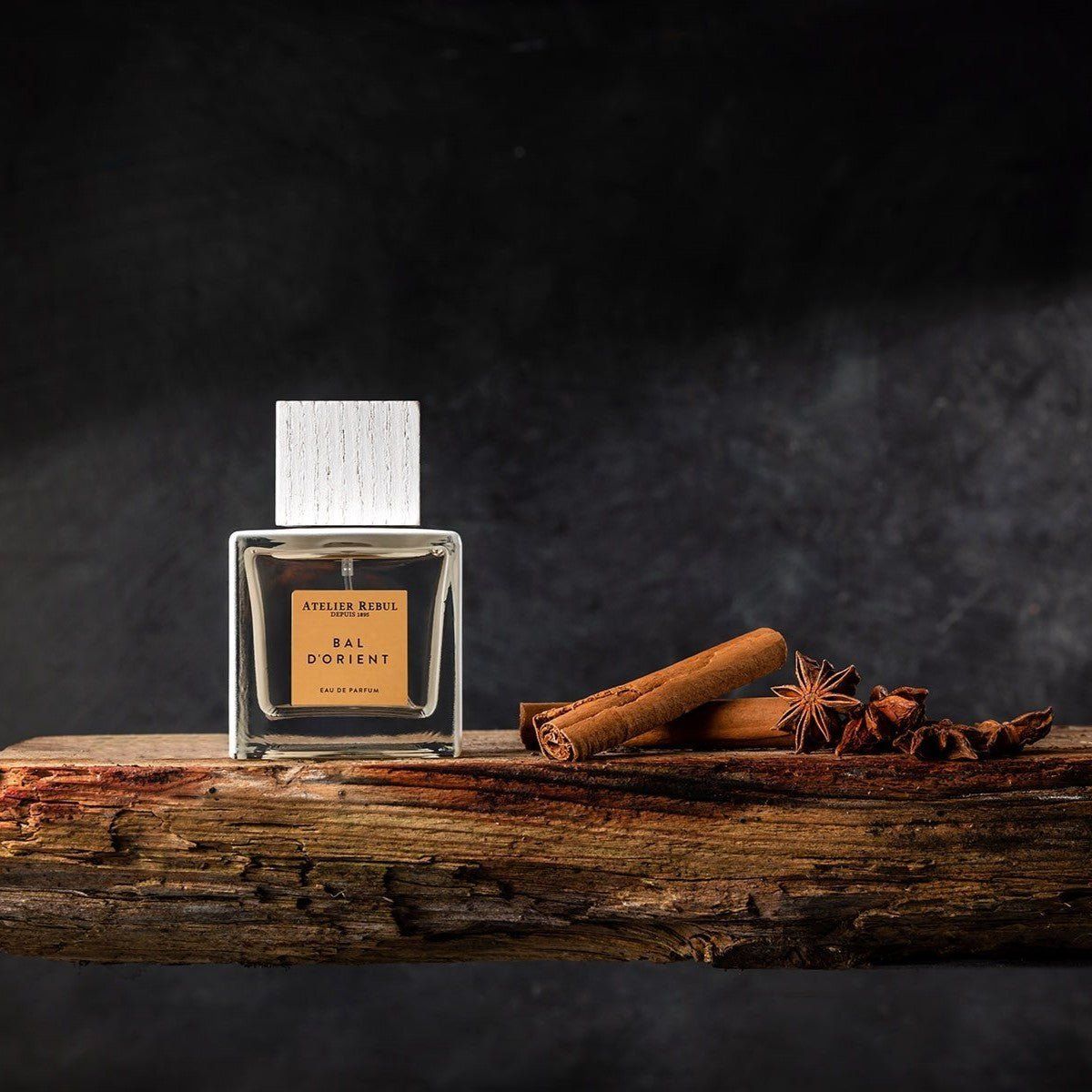 Parfümflakon auf Holz. Eckiger Flakon mit hellem Inhalt, weißer Deckel. Goldfarbenes Etikett. Zimtstangen und Sternanis.