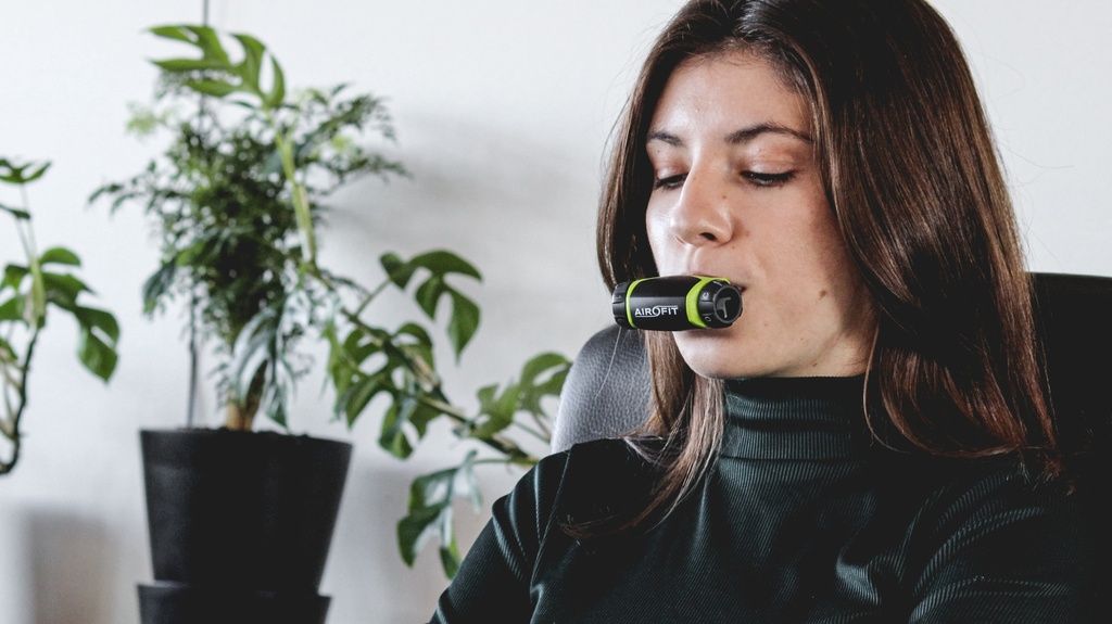 Frau trainiert mit Atemtrainer. Schwarz-grünes Gerät im Mund. Frau sitzt vor Pflanze.