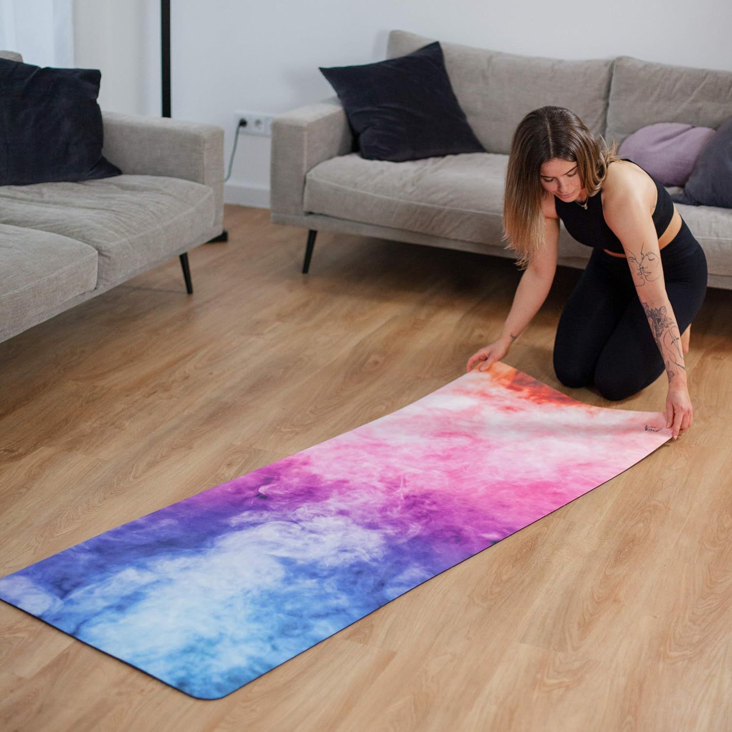 Frau rollt Yogamatte aus. Pink-blaue Matte auf Holzboden. Sofa im Hintergrund.