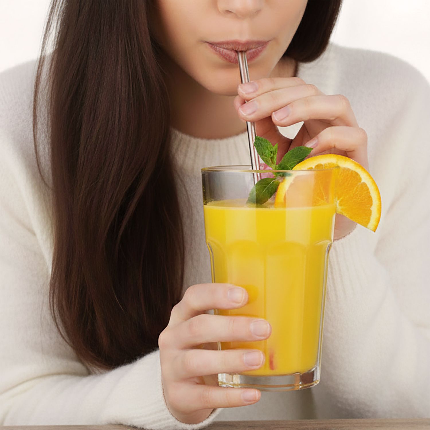 Glas mit Orangensaft, Trinkhalm, Orangenscheibe und Minze. Person trinkt.