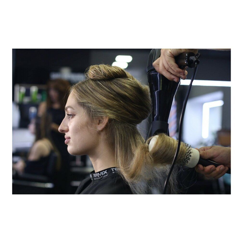 Friseur föhnt die Haare einer Frau mit einer Rundbürste. Im Hintergrund weitere Personen.