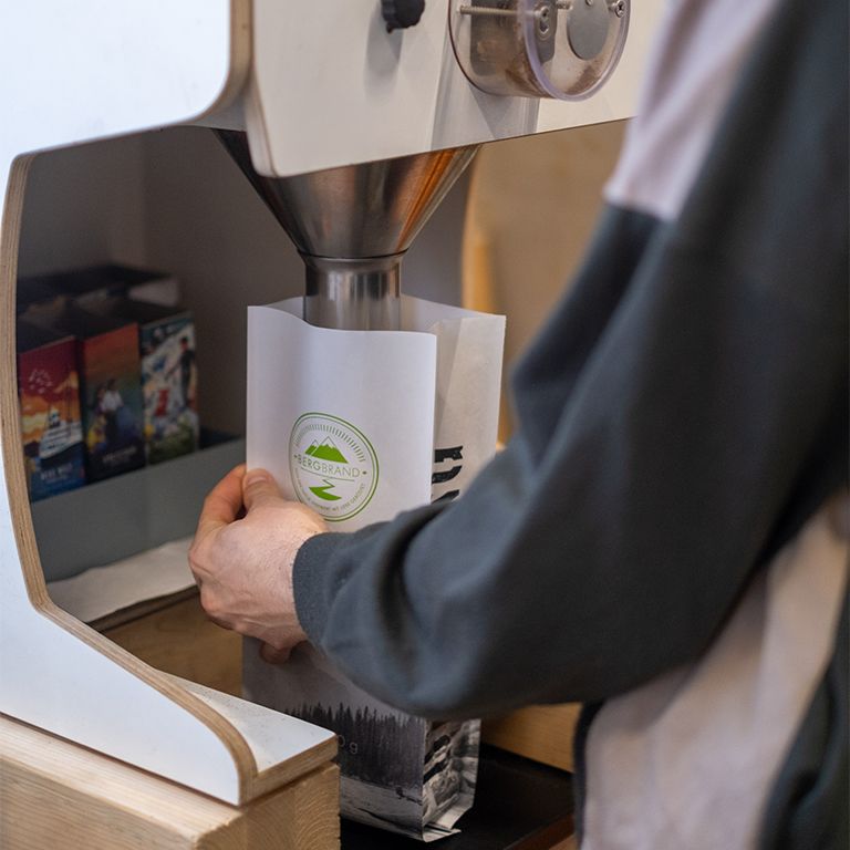 Person befüllt Kaffeebohnen-Packung. Packung mit Logo. Im Hintergrund weitere Packungen.