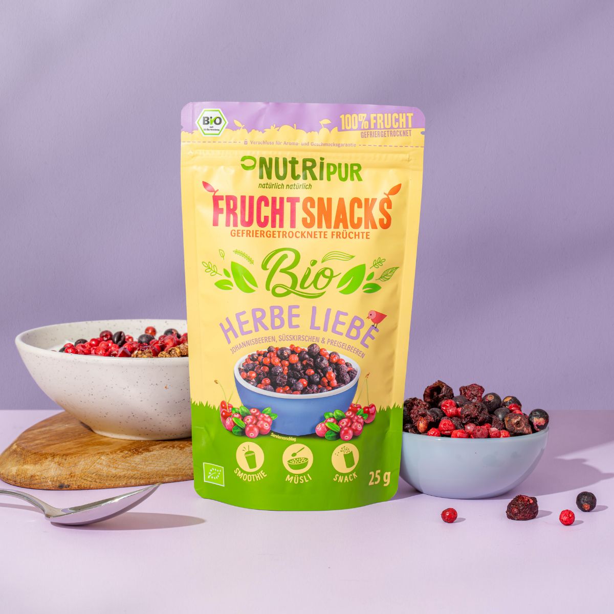 Produktverpackung und Schalen mit gefriergetrockneten Früchten auf Holzbrett. Löffel daneben. Bio-Siegel. 25g.