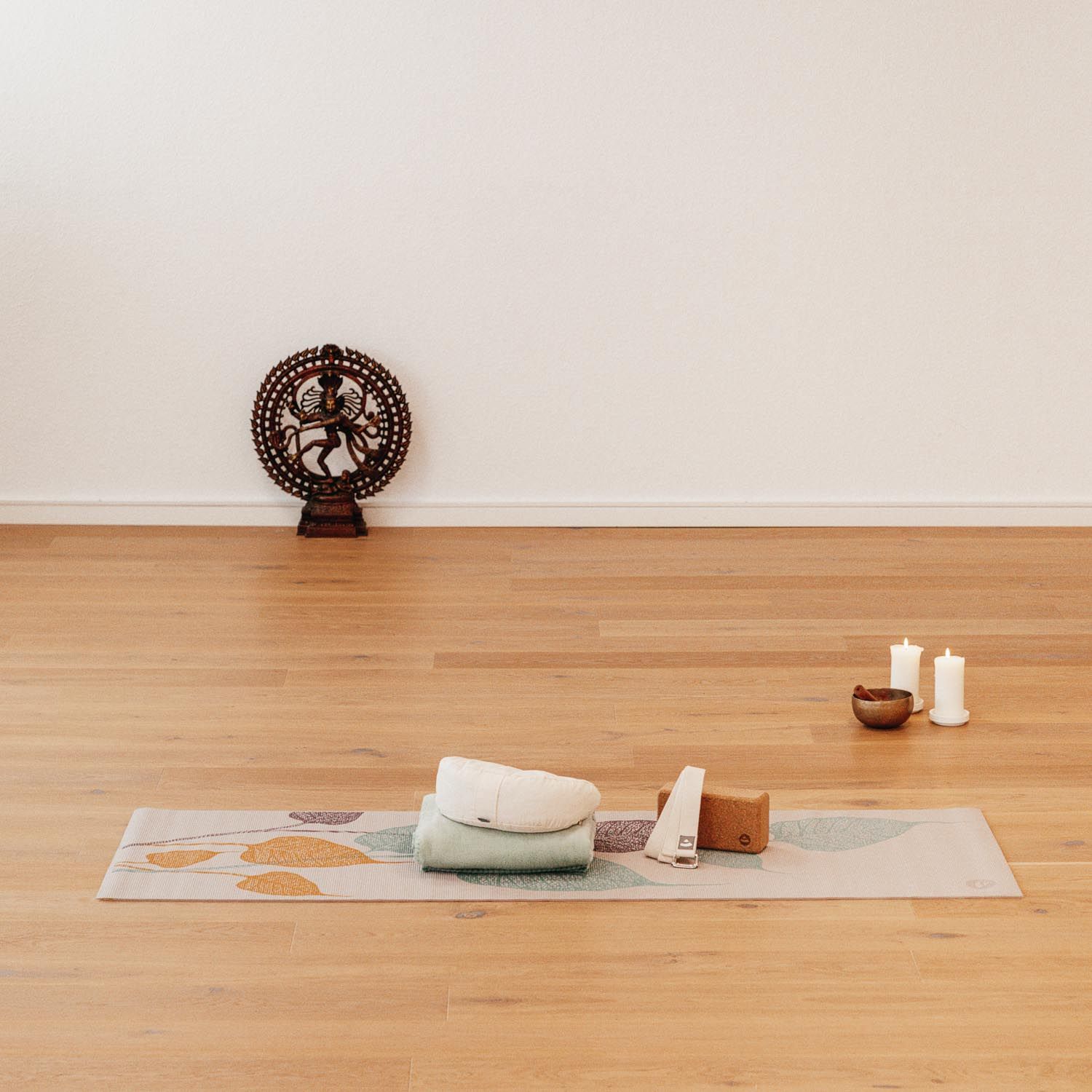 Yogamatte mit Blättermuster, umgeben von Yoga-Zubehör: Handtuch, Blöcke, Kerzen und Statue.