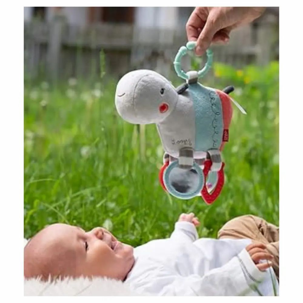 Baby liegt auf Gras, Hand hält Nilpferd-Kuscheltier. Spielzeug mit Spiegel und Ringen. Baby schaut.