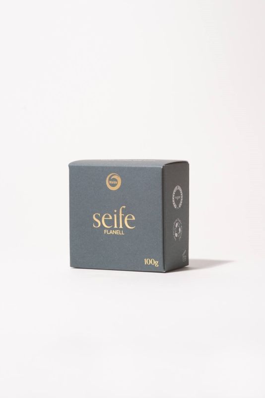 Dunkelgraue Schachtel mit goldfarbenem Schriftzug "seife FLANELL" und Logo. Aufschrift "100g".