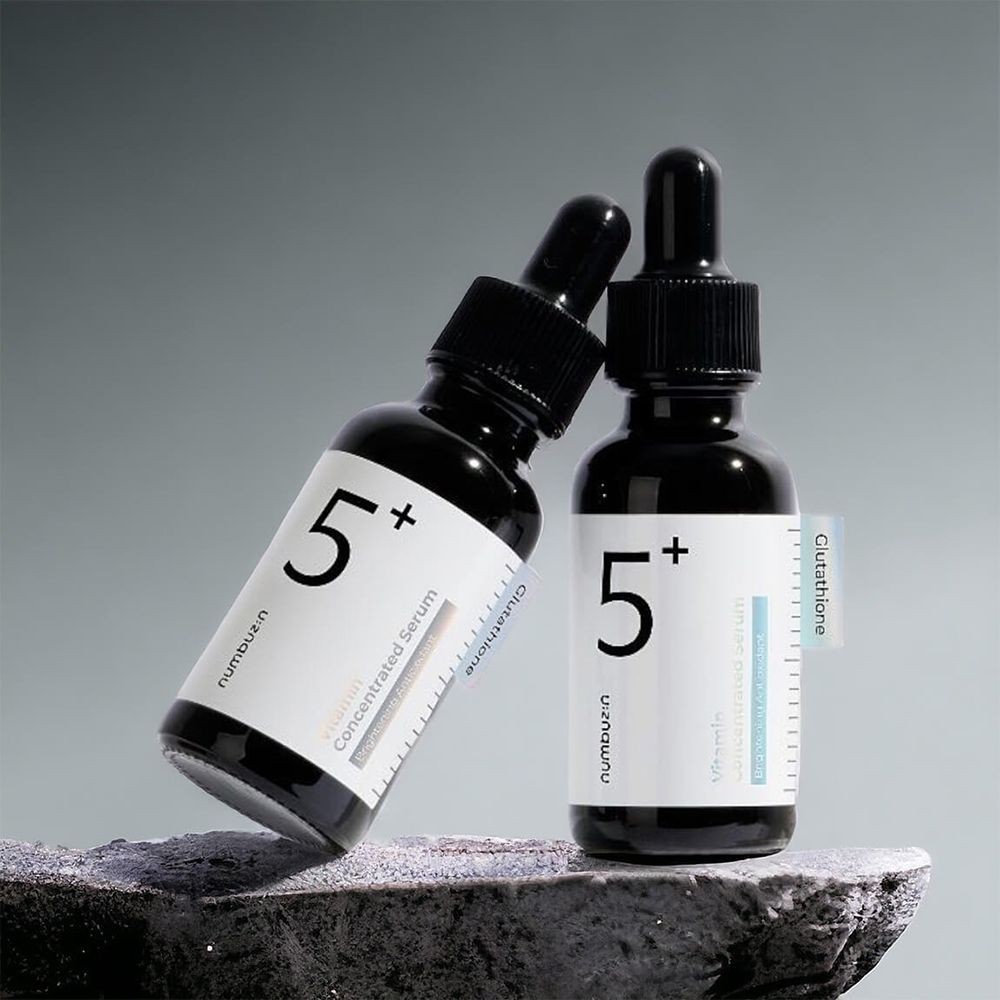 Zwei schwarze Flaschen mit weißem Etikett. Aufschrift "5+" und "numbuzin". Vitamin-Serum. Mit Tropfern. Auf Stein platziert.