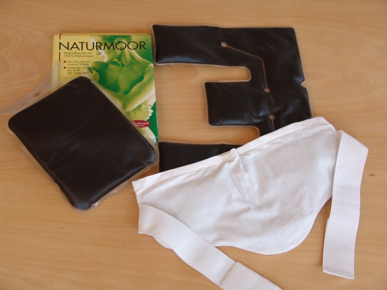 Verschiedene Teile des Beckenwärmers. Schwarze Kissen, weißer Gurt und Verpackung mit Text: NATURMOOR.