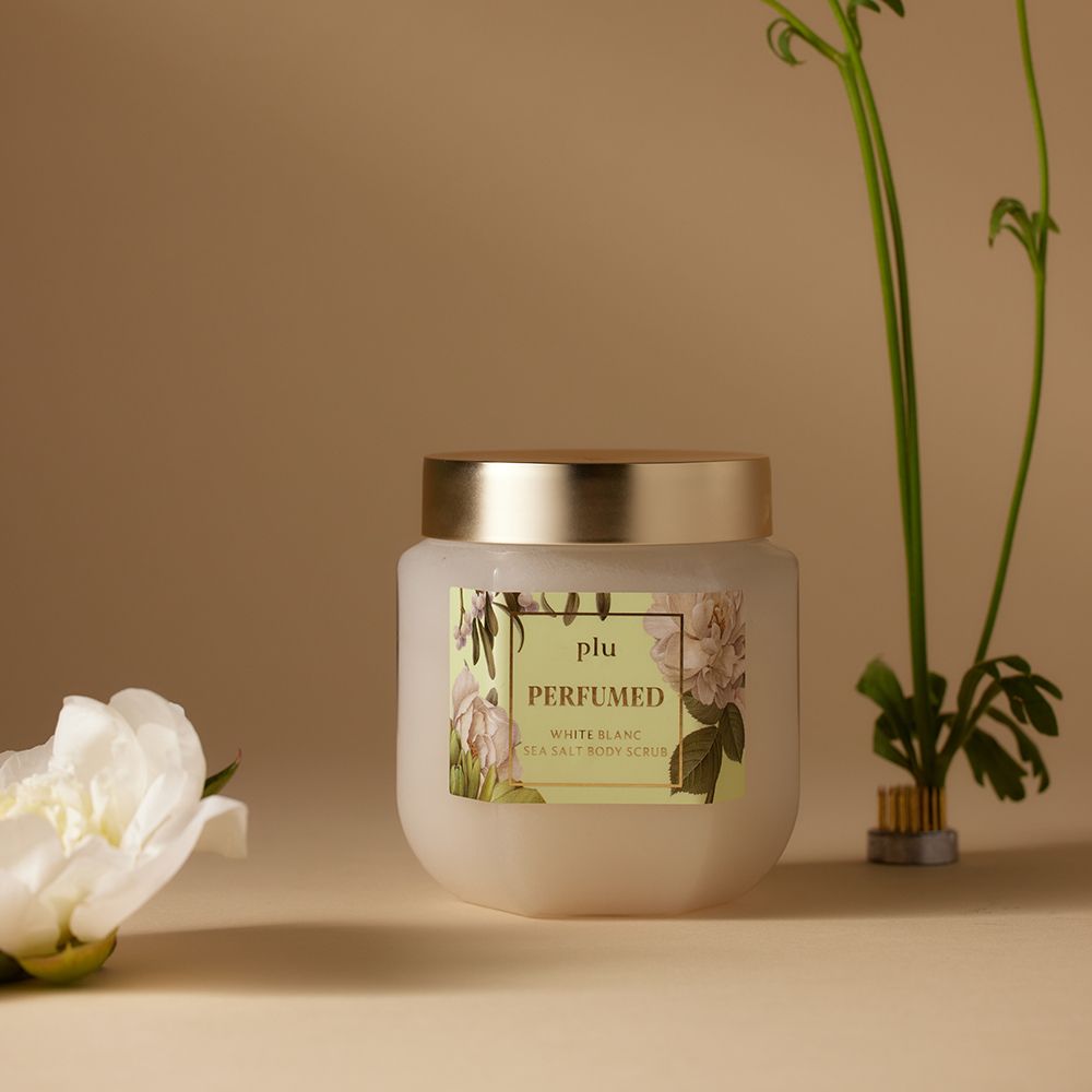 Glasbehälter mit goldfarbenem Deckel und Etikett. Aufschrift: PLU PERFUMED White Blanc Sea Salt Body Scrub. Floral-Dekor. Beige Hintergrund.
