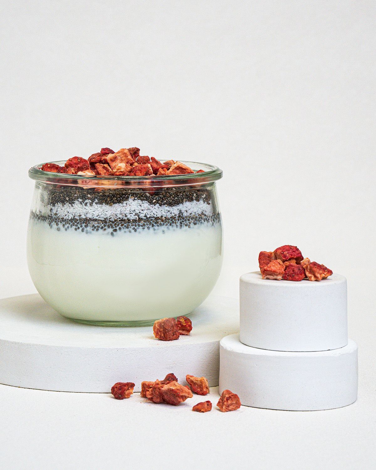 Glas mit Joghurt, Chia-Samen und gefriergetrockneten Erdbeerstücken. Erdbeerstücke auf Podest. Produktpräsentation.