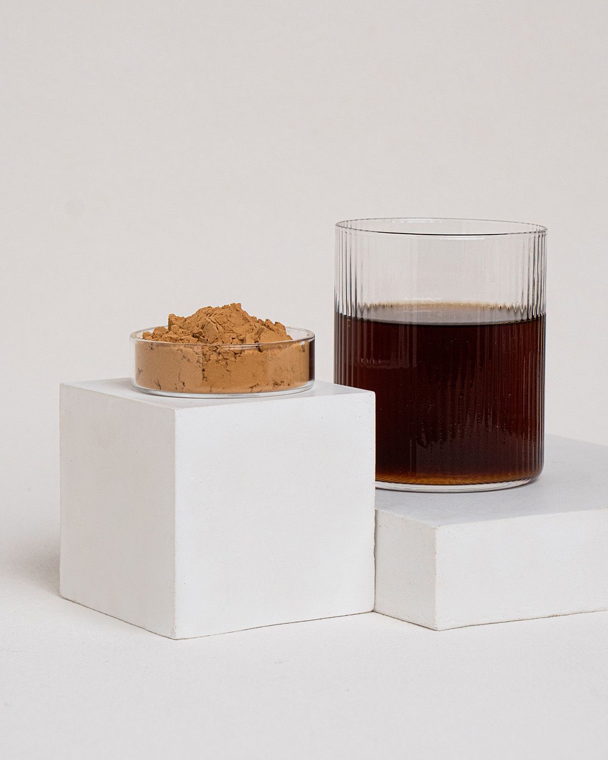 Braunes Pulver in einer kleinen Glasschale. Daneben ein Glas mit dunkler Flüssigkeit, auf weißen Blöcken platziert.