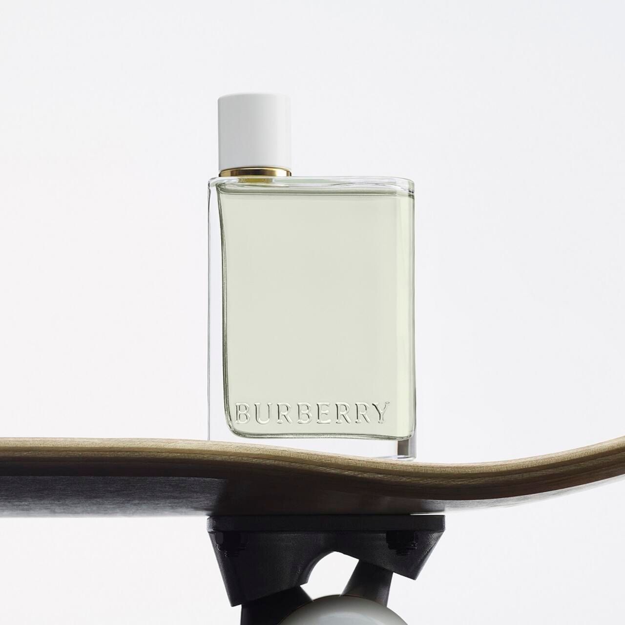 Burberry Her Eau de Toilette Flakon auf einem Skateboard. Weißer Deckel, goldener Kragen, transparente Flüssigkeit. Logo.