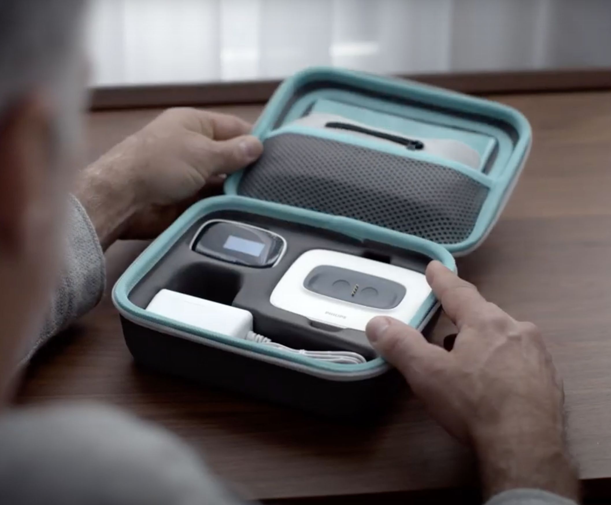 Geöffnete Transportbox mit Schlafpositionstrainer und Zubehör. Hände halten die Box. Philips-Logo sichtbar.