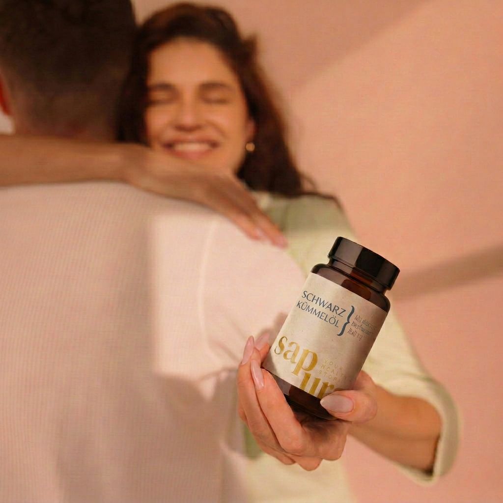 Frau umarmt Mann und hält eine braune Flasche mit Kapseln. Aufschrift: SCHWARZKÜMMELÖL, Kaltgepresst & Naturbelassen, sapura.