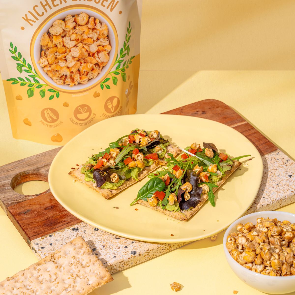 Gelber Teller mit zwei Knäckebroten, belegt mit Avocado, Gemüse und Flakes. Im Hintergrund eine Packung Knusperflakes Kichererbsen und eine Schüssel mit Flakes.