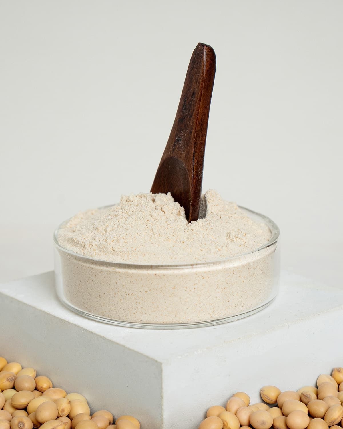 Glasbehälter mit buXtrade Nattokinase Pulver und einem Holzspatel. Sojabohnen im Vordergrund.