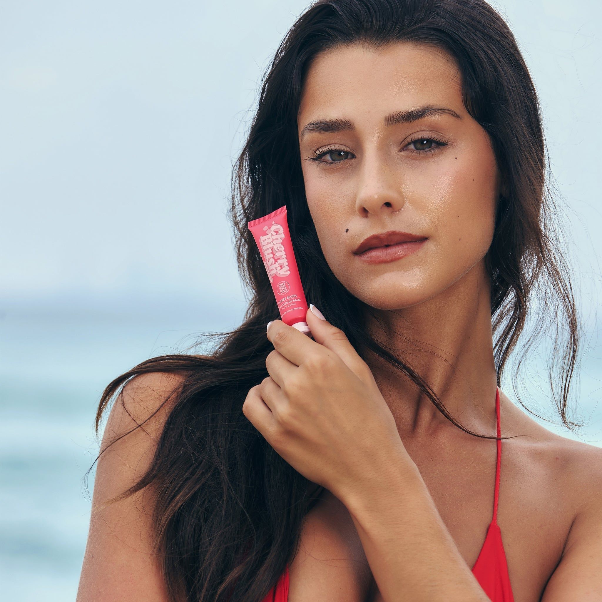 Frau hält Tube 'Cherry Blush'. Produktname und Marke sichtbar. Rotes Oberteil. Strandhintergrund.