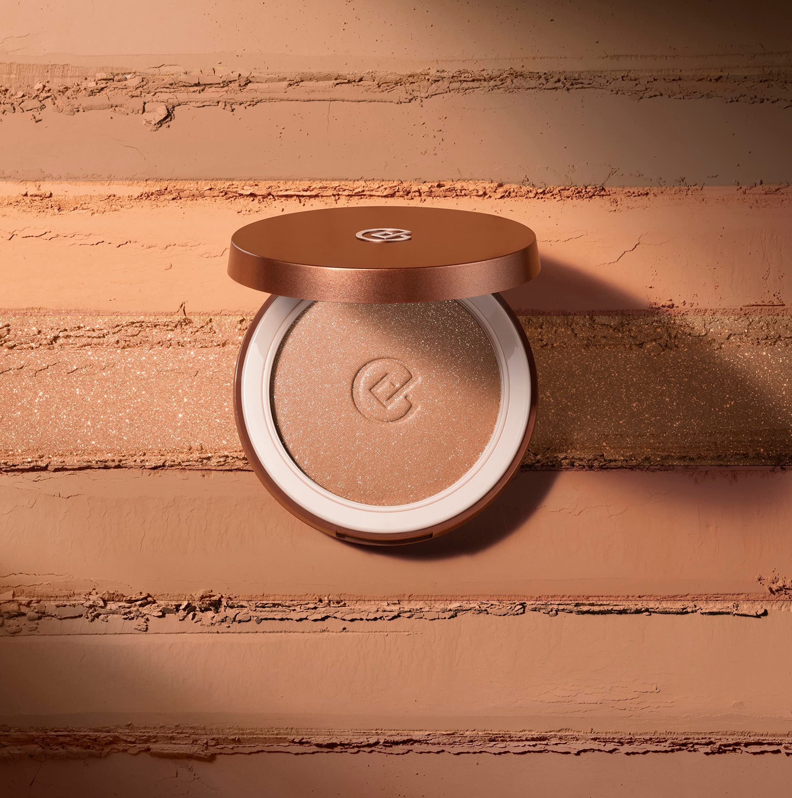 Bronzer mit geöffnetem Deckel. Helle Puderfarbe. Geprägtes Logo. Auf gestreiftem, farbigen Hintergrund.