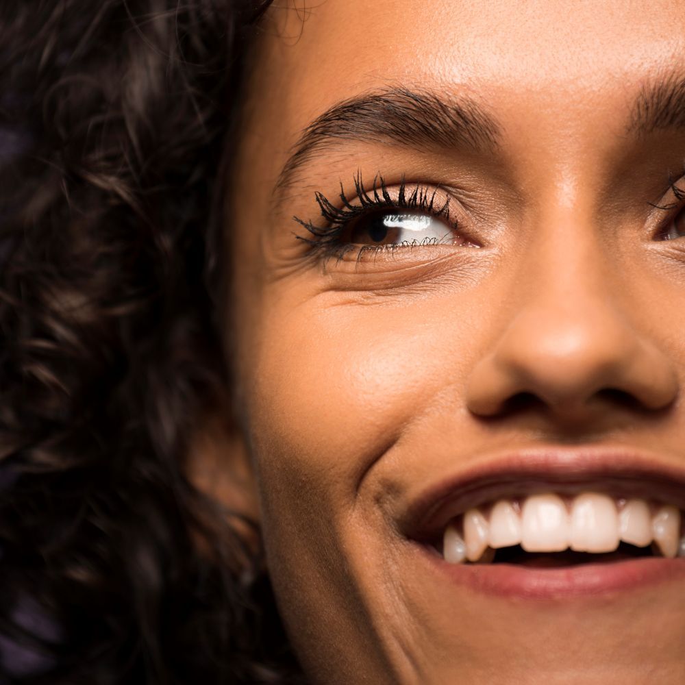Nahaufnahme eines lächelnden Gesichts mit getuschten Wimpern. Hautton, Augenbrauen und Augen sind sichtbar.