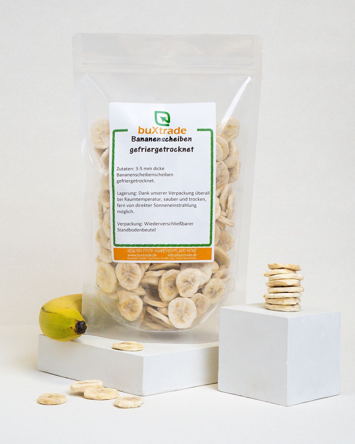 Verpackung mit gefriergetrockneten Bananenscheiben, daneben eine Banane und gestapelte Scheiben.