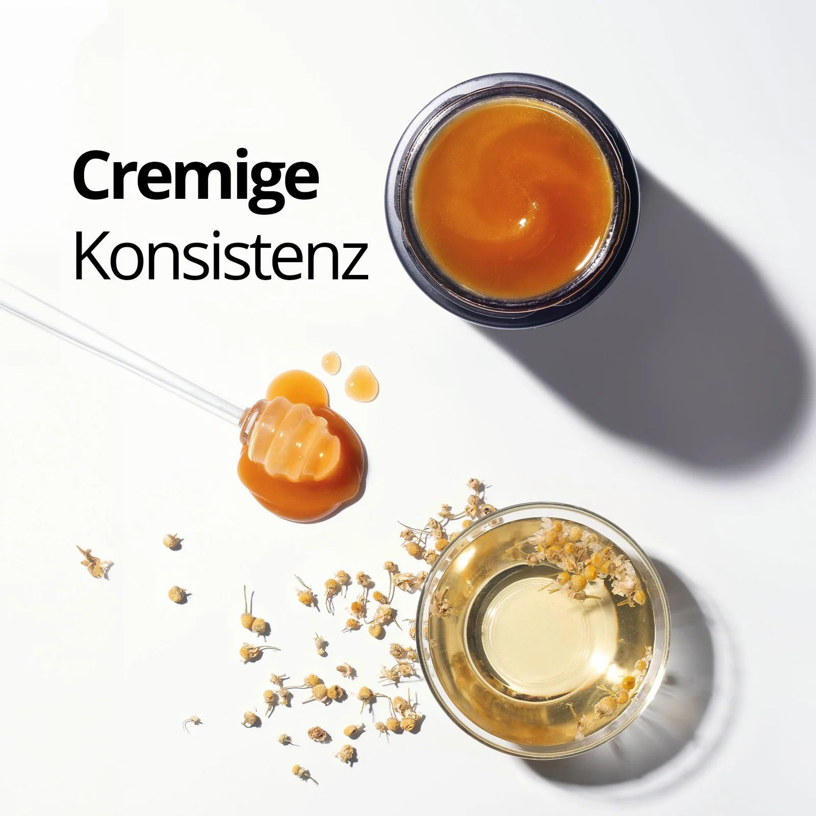 Offenes Honigglas mit cremiger Konsistenz, Honigwabe und Kamillenblüten. Text: Cremige Konsistenz.