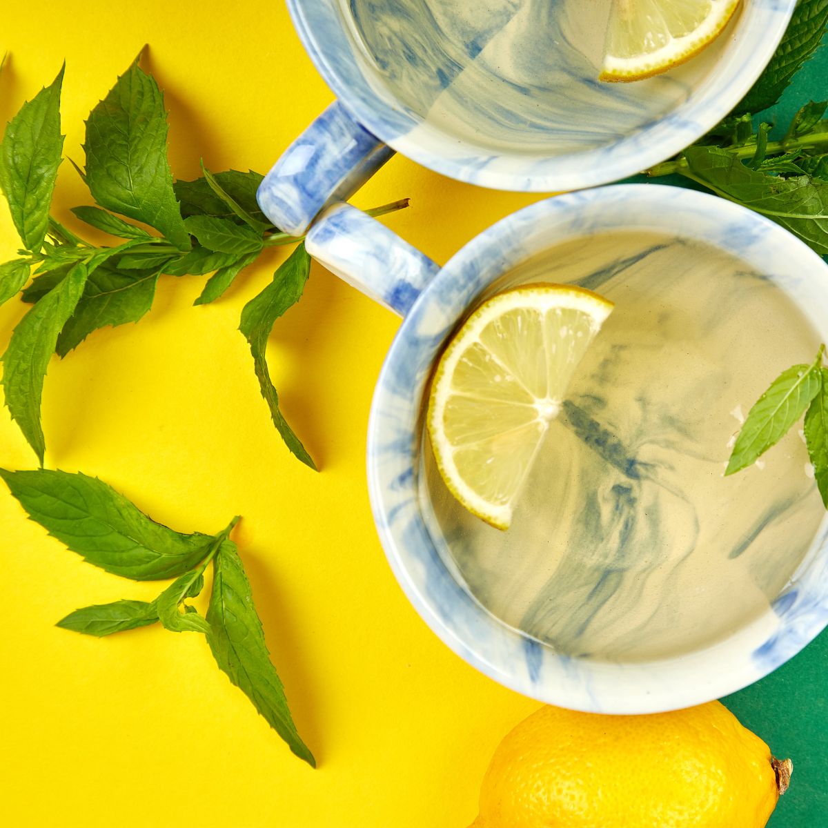 Zwei Tassen Tee mit Zitronenscheiben, umgeben von Minzblättern und einer Zitrone. Gelber und grüner Hintergrund.