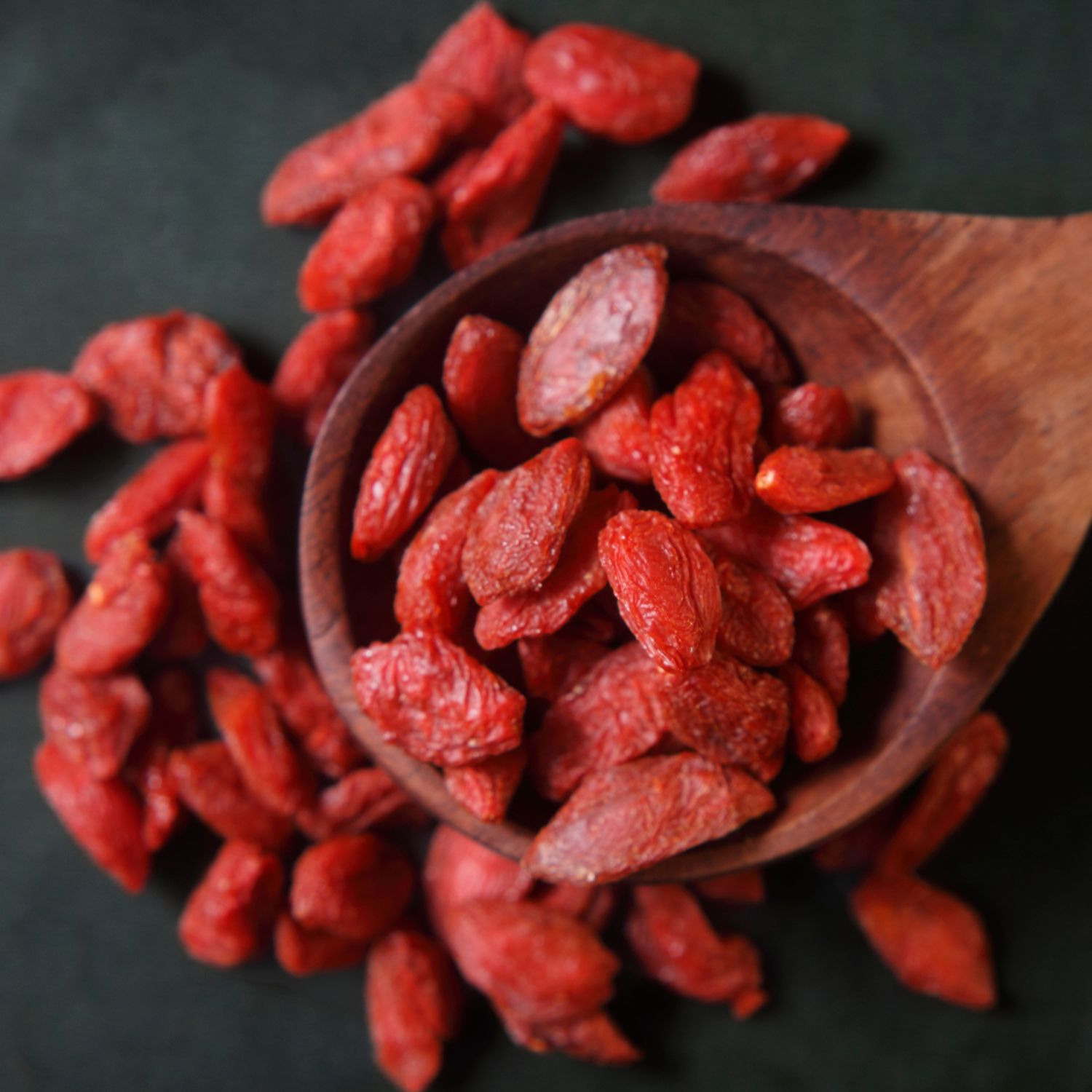 Goji-Beeren auf dunklem Untergrund. Einige Beeren liegen in einem hölzernen Löffel. Rote Farbe.