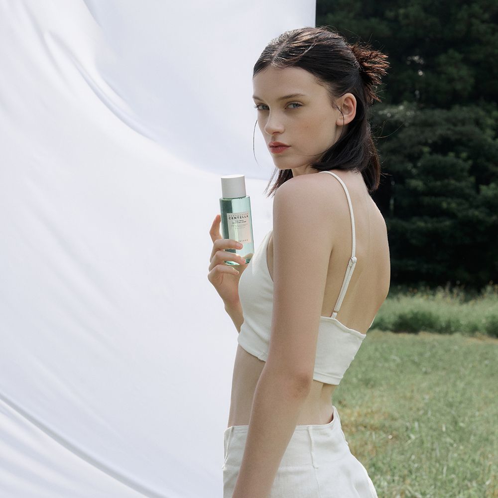 Junge Frau hält eine Flasche mit grünlicher Flüssigkeit. Sie trägt ein weißes Top und Hose. Hintergrund: Natur, weißes Tuch.