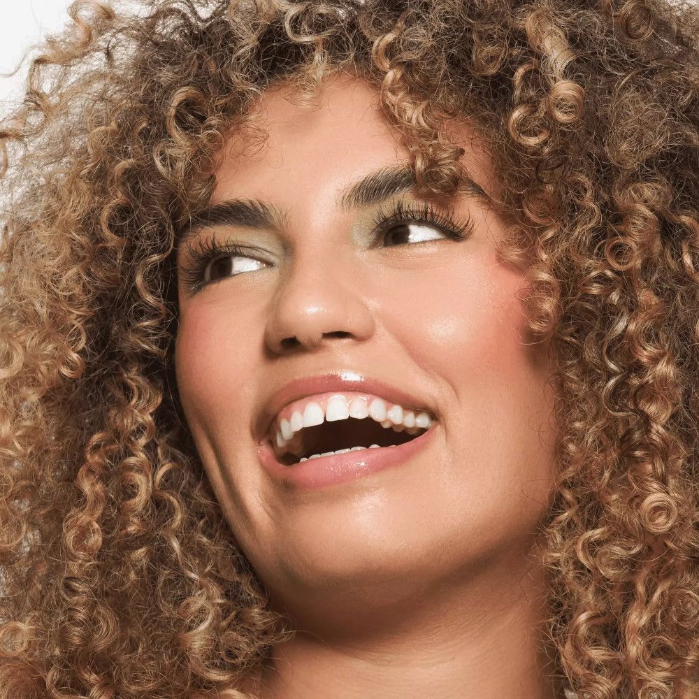 Porträt einer lächelnden Frau mit lockigem Haar. Sie trägt grünen Lidschatten und glänzenden Lippenbalsam.