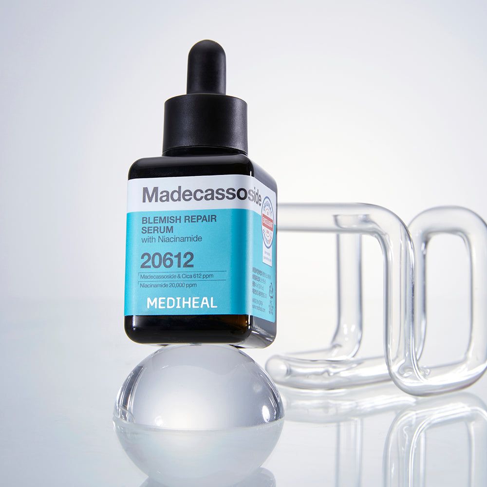 Serumflasche auf einer Kugel und Glasobjekt. Schwarzer Deckel, quadratische Flasche mit blauem Etikett. Text: Madecassoside Blemish Repair Serum, 20612, MEDIHEAL.
