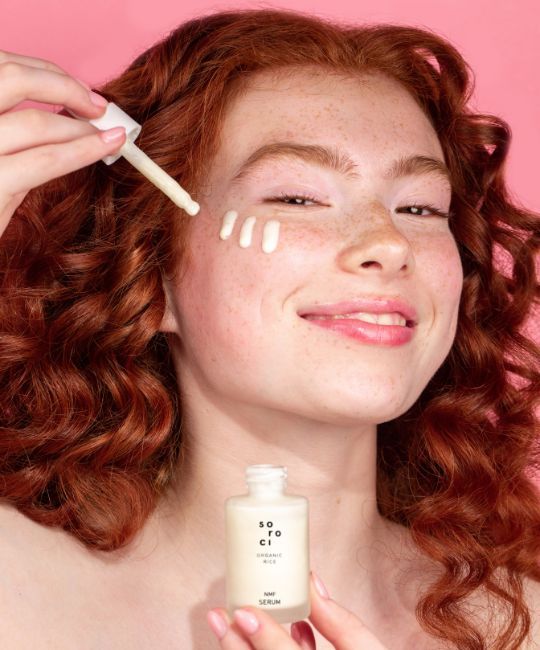 Frau mit rotem Haar, Serum auftragend. Flasche mit weißem Deckel und Aufschrift. Serum auf der Haut. Rosa Hintergrund.