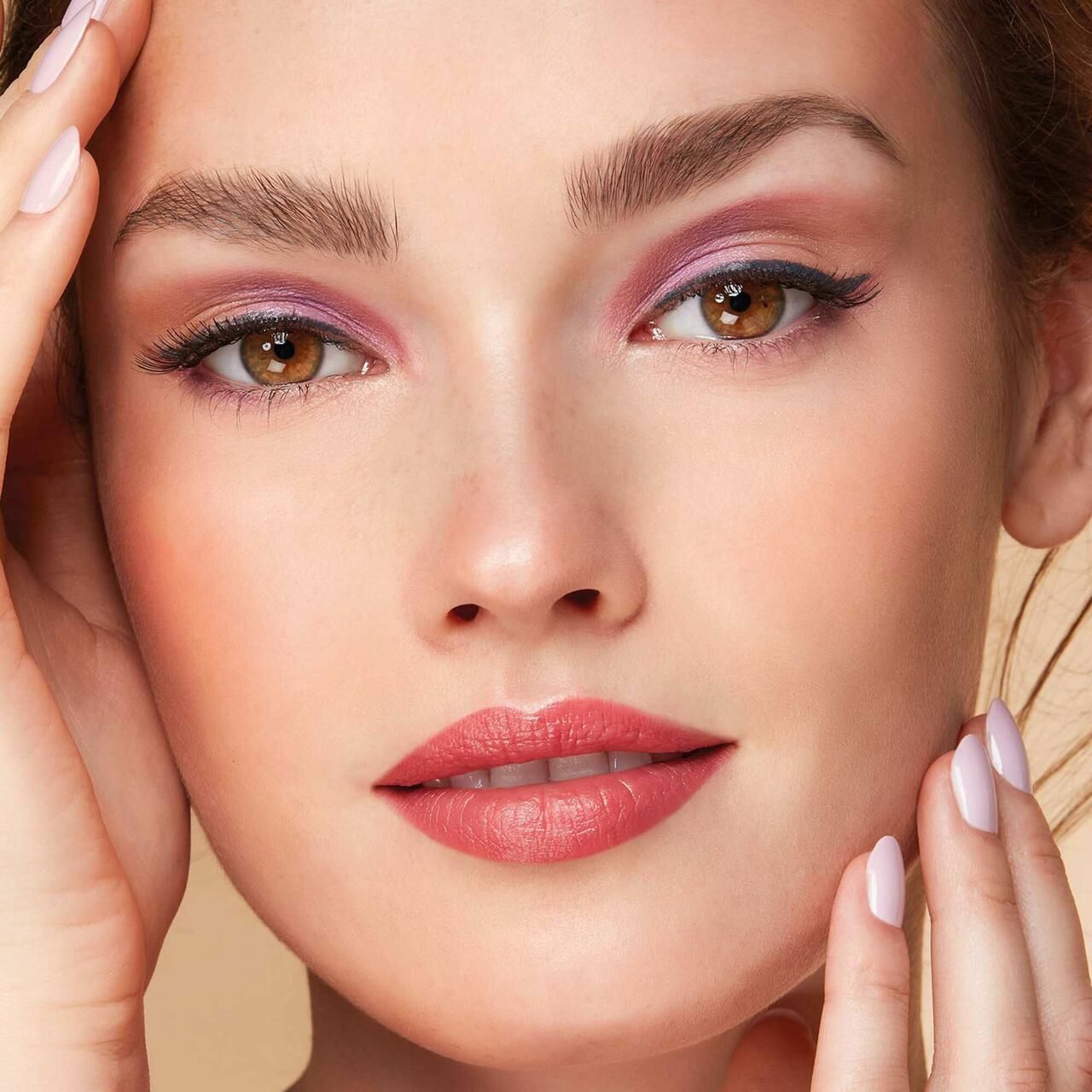 Nahaufnahme eines Gesichts mit pinkfarbenem Eyeshadow. Hände berühren das Gesicht. Lippen rot geschminkt.