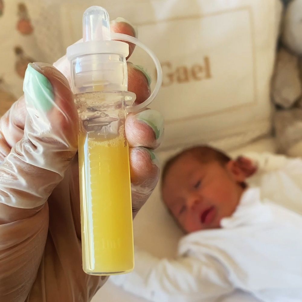 Transparenter Behälter mit Flüssigkeit, gehalten von einer Hand. Baby im Hintergrund.