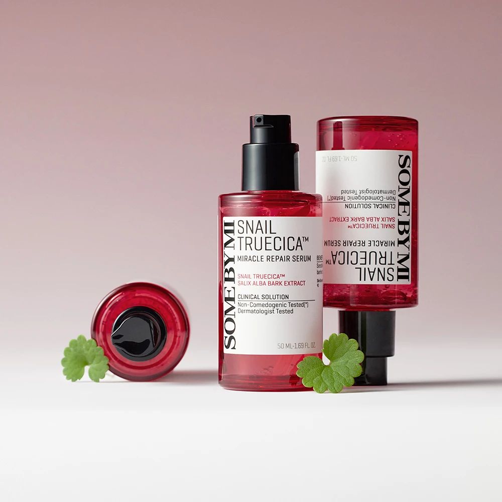 Zwei Serumflaschen mit schwarzem Pumpverschluss und eine Kappe. Produktetiketten sind sichtbar. Grüne Blätter liegen daneben.