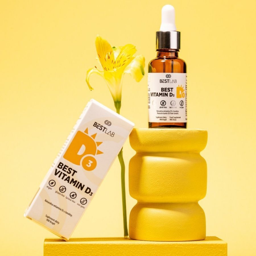 Braune Glasflasche und Karton mit Best Vitamin D3. Auf gelbem Podest mit Blume. Vegan, glutenfrei, laktosefrei, ohne Gentechnik.